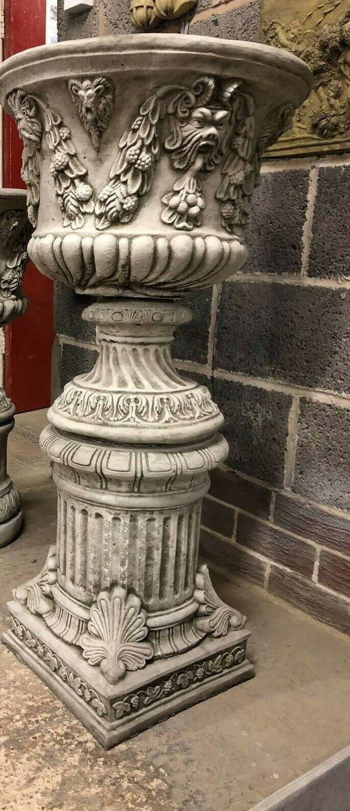 Tall stone Bacchus urn planter on fluted plinth, featuring intricate foliage and face carvings for a decorative garden focal point.