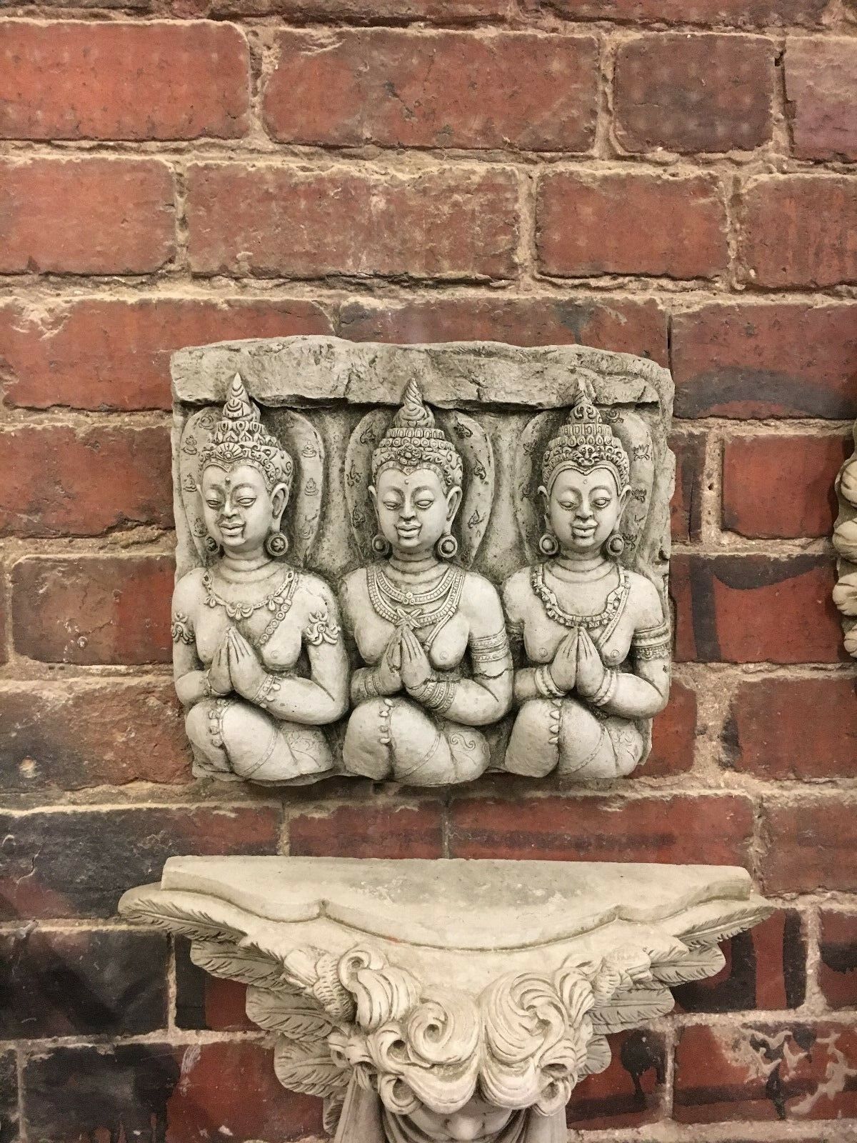 Limestone Buddha wall plaque mounted on brick wall, showcasing three serene figures. Weather-resistant garden decoration with antique finish.