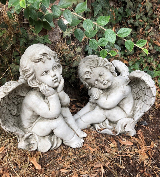 Stone winged baby angels garden ornaments pair resting on forest floor. Ideal for outdoor decor in flowerbeds or memorial garden settings.
