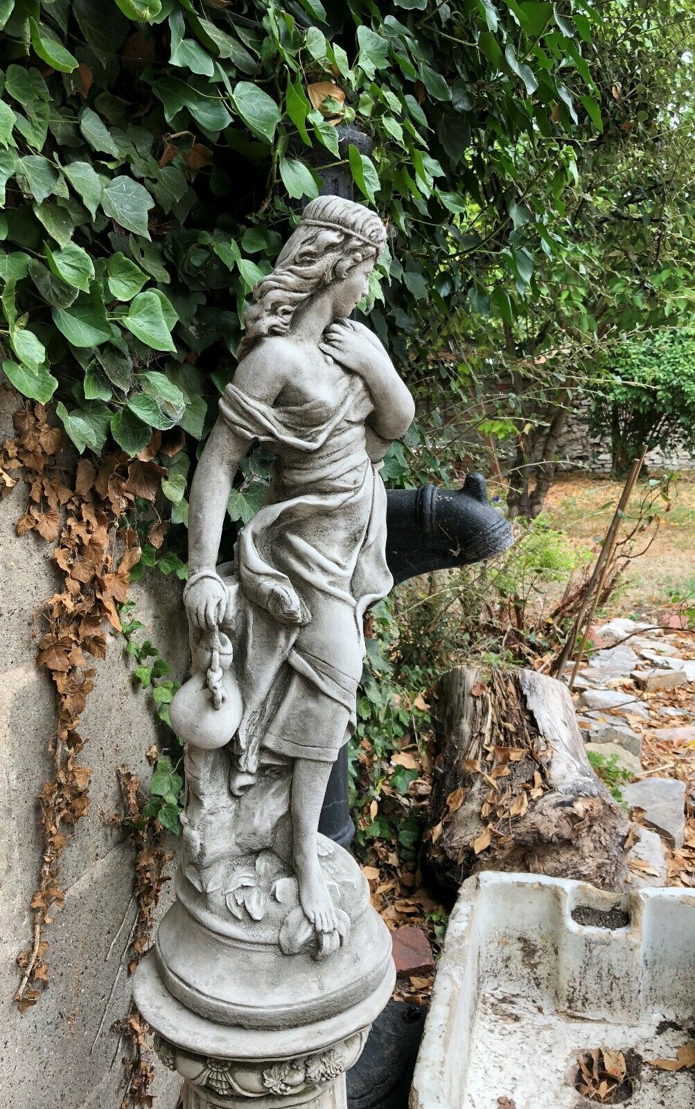 Side view of Garden Stone Statue - Jug Lady Plinth in antique grey finish, placed in a rustic garden setting with ivy-covered wall.