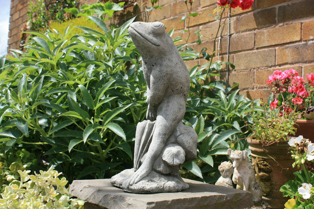 Stone Frog Garden Ornament with Umbrella on a garden pedestal, surrounded by lush greenery and flowers, ideal for patios or lawns.