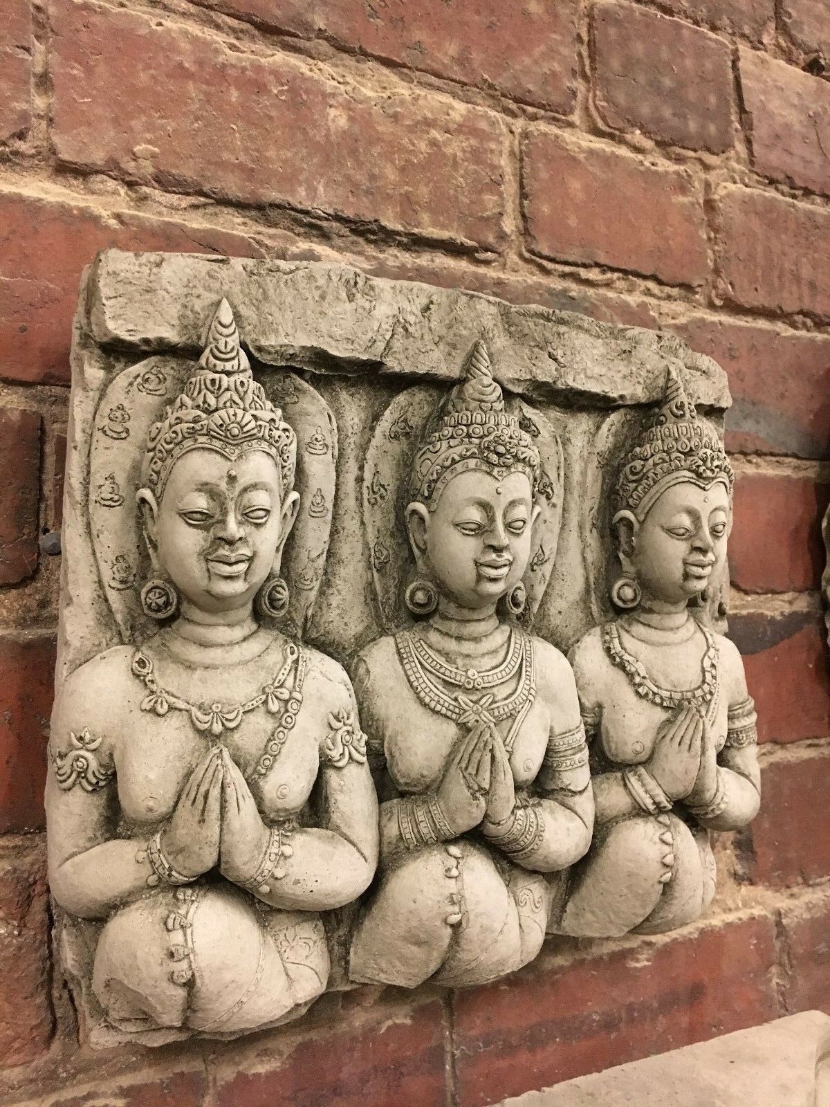 Angled view of three Buddha figures on limestone wall plaque. Durable garden ornament with intricate detailing and spiritual charm.