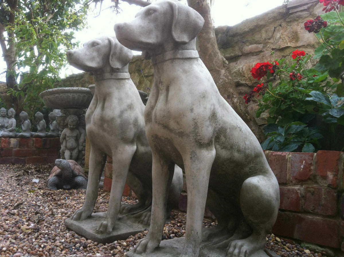 Durable antique grey stone pointer dog statues displayed in a gravel garden with red flowers and decorative garden ornaments.