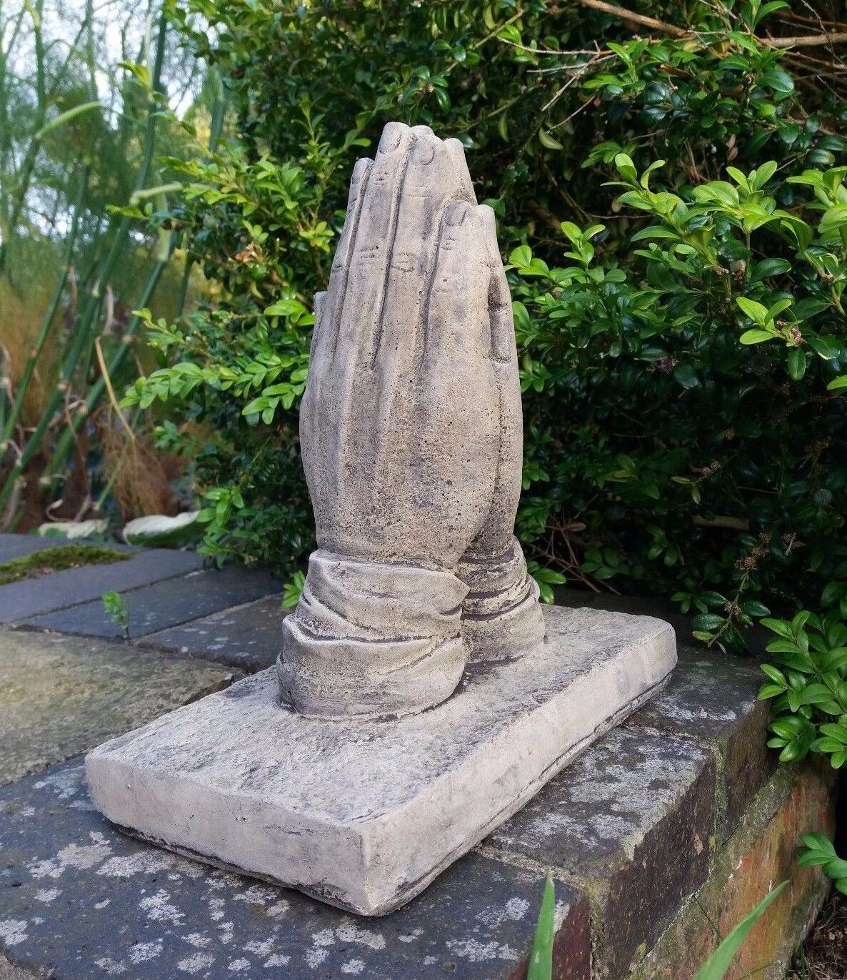 Stone prayer hands ornament on rectangular base, handcrafted for memorial garden decor. Frostproof limestone sculpture with aged finish.