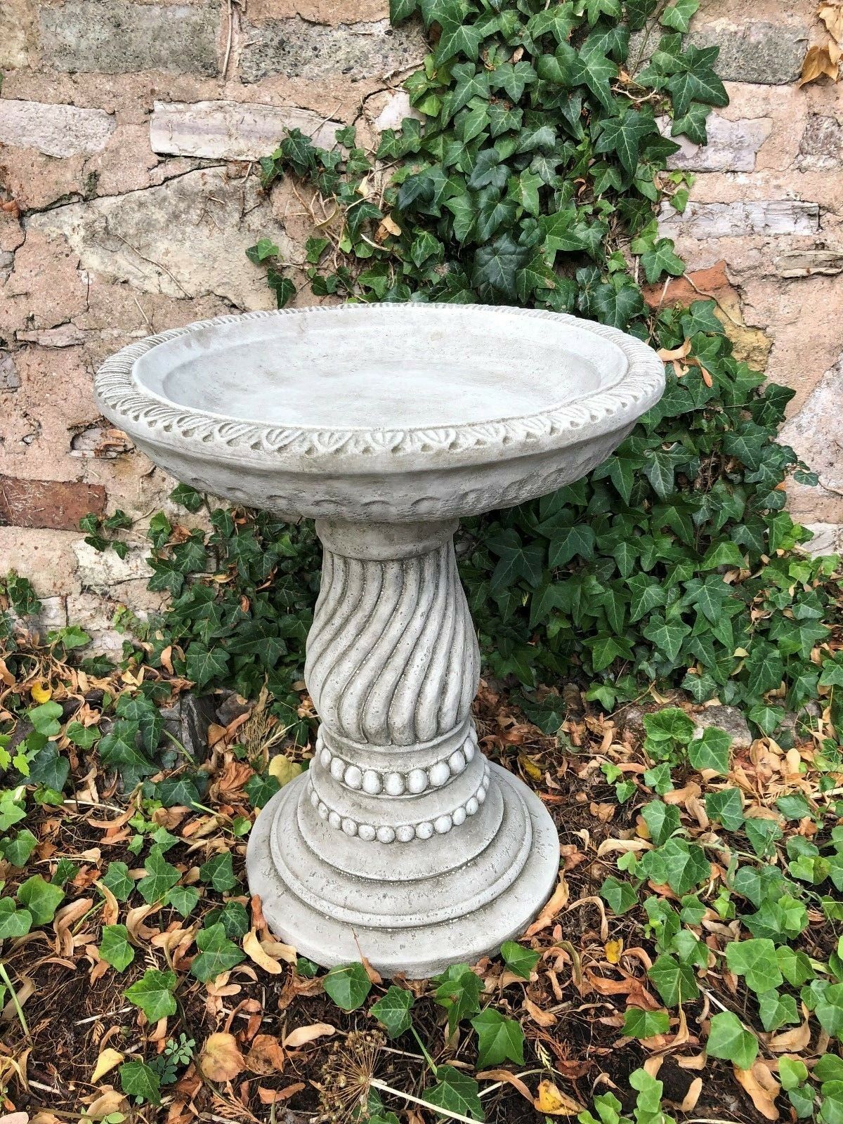Stone garden bird bath with twist base design placed against ivy-covered stone wall. Durable limestone ideal for outdoor bird feeding.
