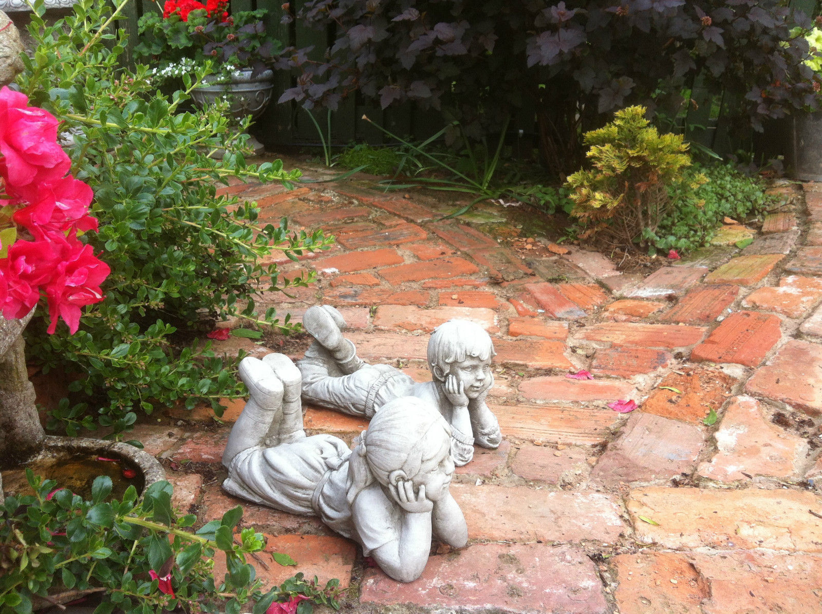 Pair of stone garden ornaments - boy and girl laying on brick path under blooming flowers. Durable and elegant garden sculpture set.