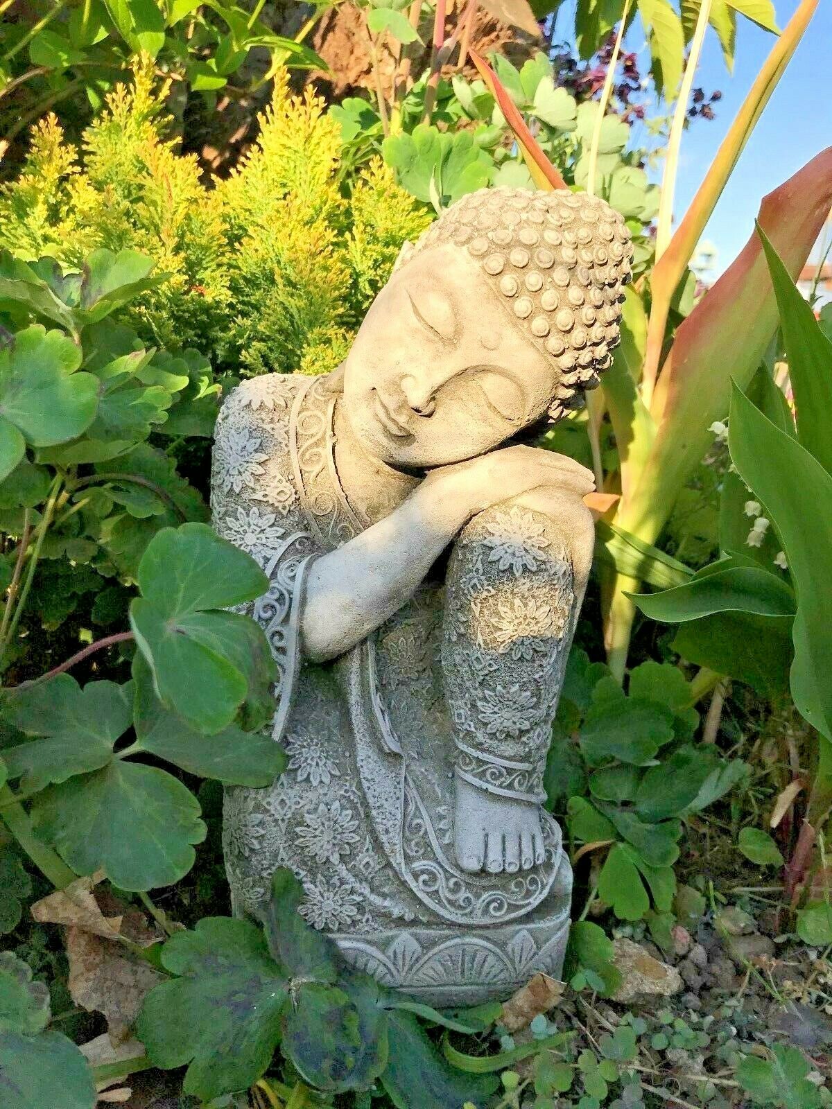 Sleeping Buddha statue in limestone finish resting peacefully in a lush garden, ideal as a stone garden ornament for outdoor zen decor.