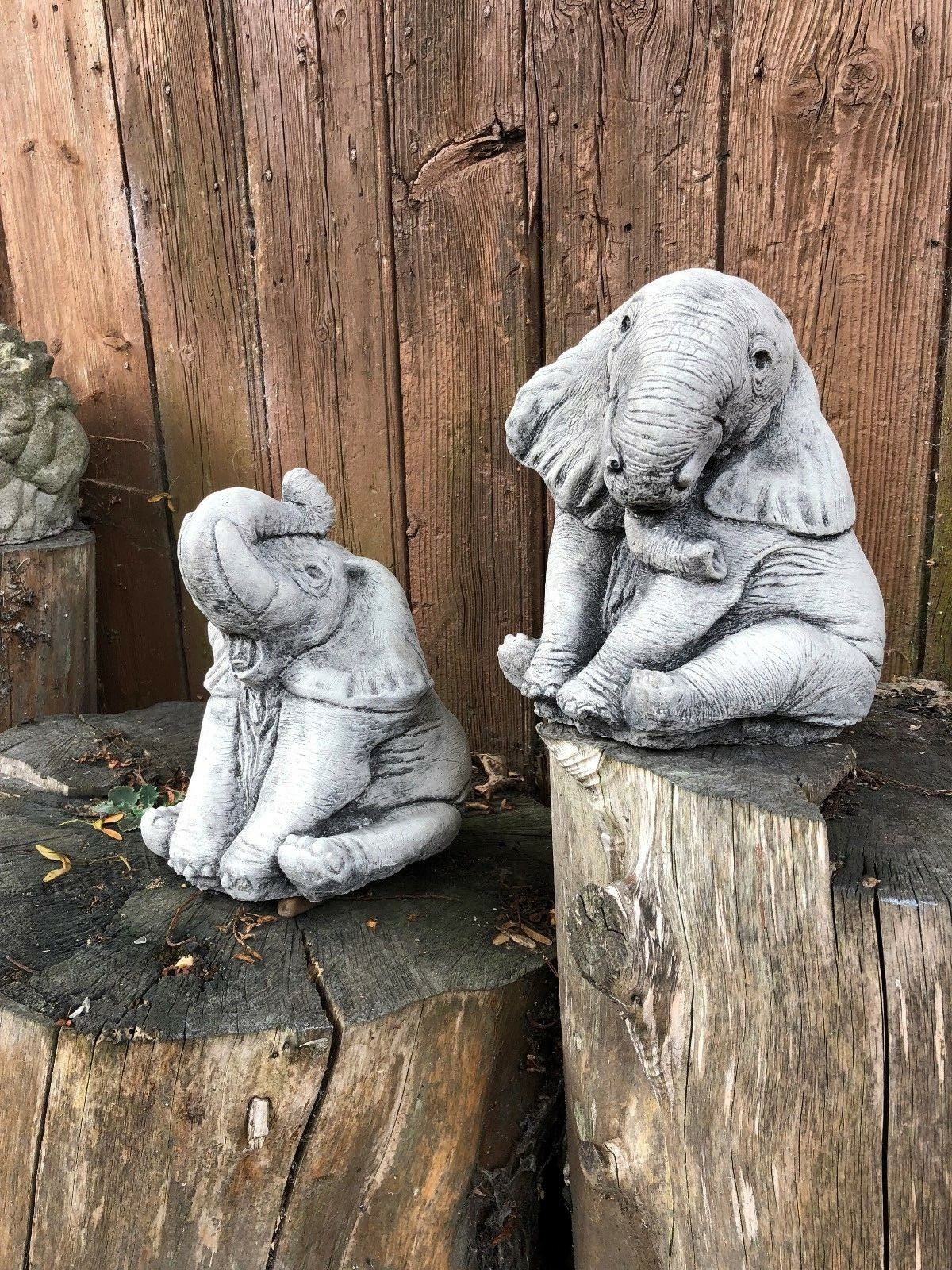 Pair of baby elephant ornaments in antique grey stone finish, displayed on tree stumps for garden decor. Ideal for outdoor lawn sculpture accents.