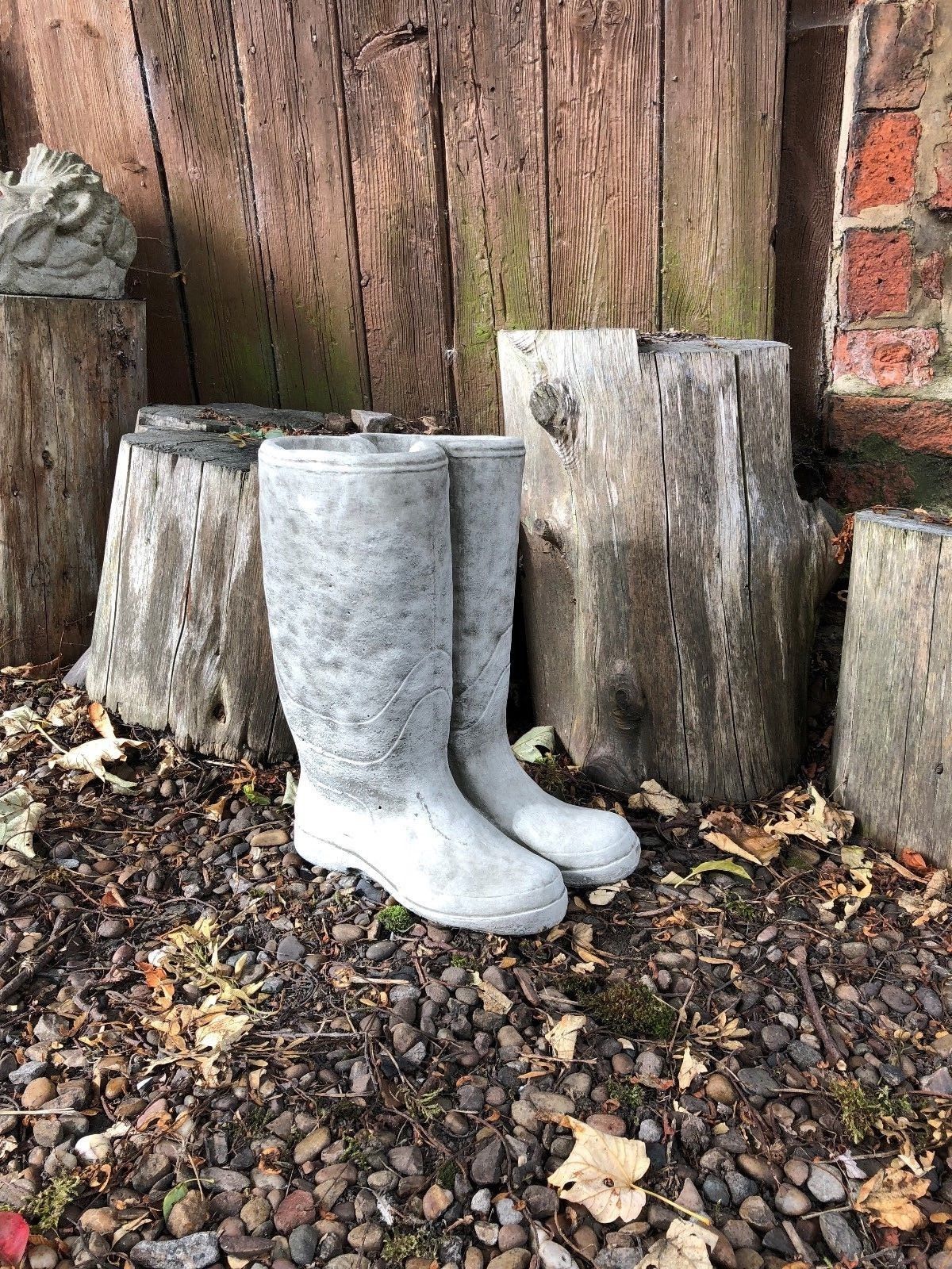 Stone Wellington Boots Planter with antique finish, placed on pebbled garden ground beside rustic wooden logs. Durable limestone garden decor.