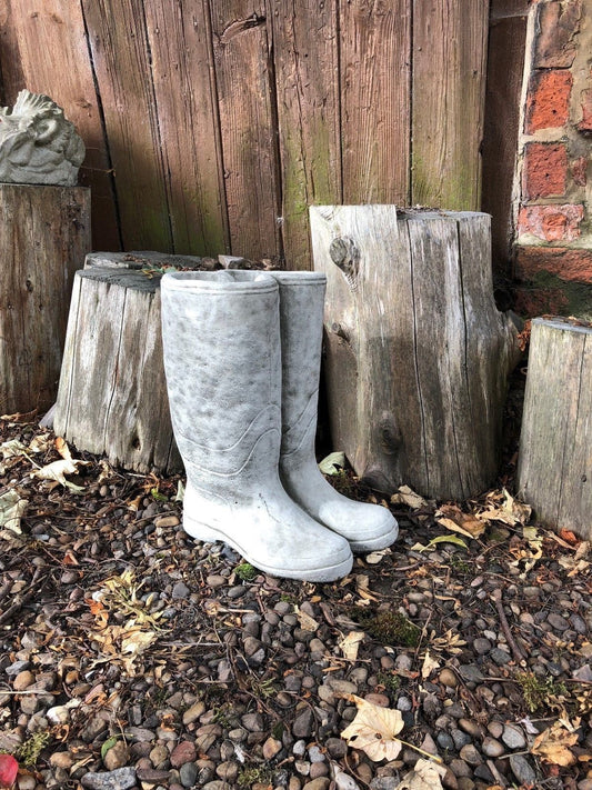 Stone Wellington Boots Planter with antique finish, placed on pebbled garden ground beside rustic wooden logs. Durable limestone garden decor.