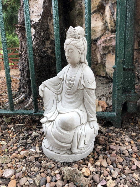 Kuan Yin garden statue in stone, seated on lotus base near green iron fence. Durable 18" limestone sculpture for outdoor garden décor.