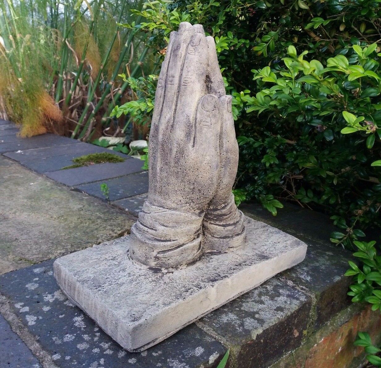 Handcrafted stone prayer hands statue on patio wall, ideal for outdoor memorial gardens. Made from frostproof crushed limestone.