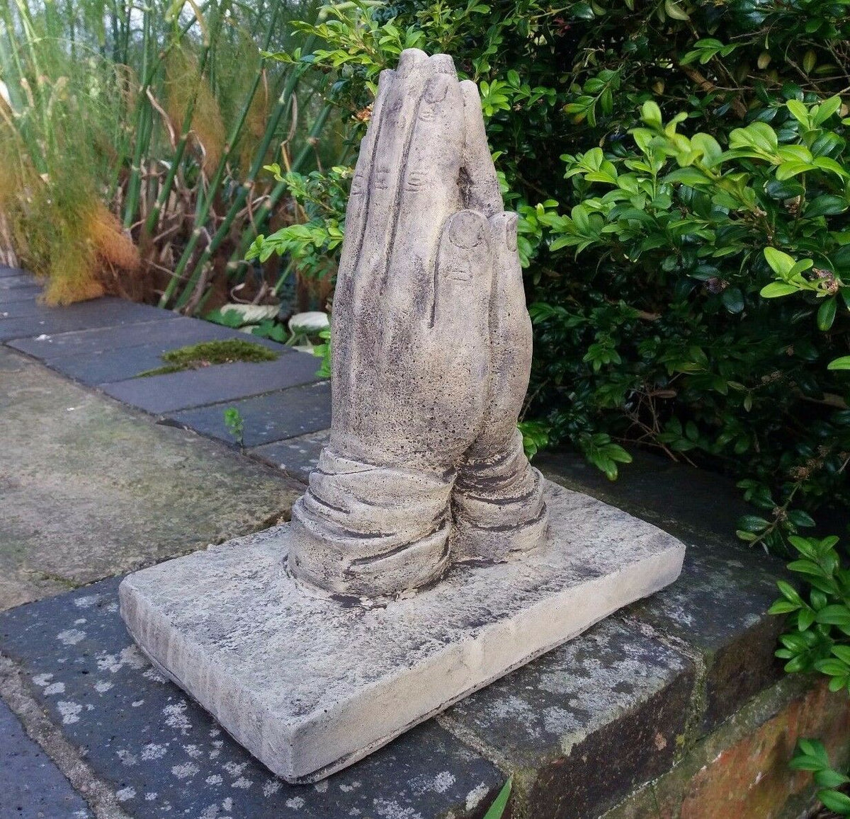 Handcrafted stone prayer hands statue on patio wall, ideal for outdoor memorial gardens. Made from frostproof crushed limestone.