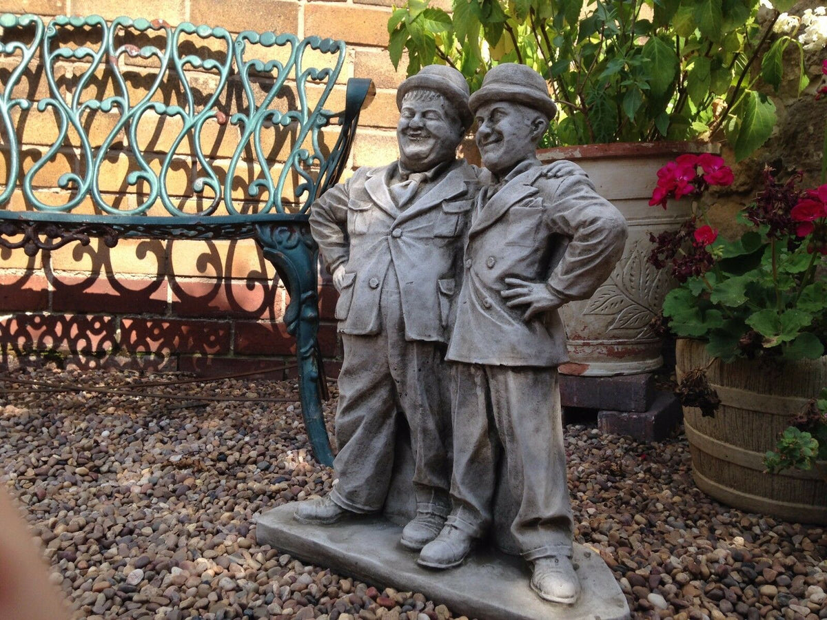Laurel & Hardy garden statue in antique stone finish, placed on gravel near a vintage bench and potted plants for outdoor decor.