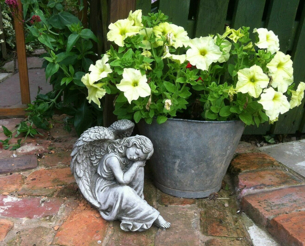 Winged Angel Ornament in antique grey stone finish placed beside a metal flower pot on a brick patio. Ideal for garden or patio decor.
