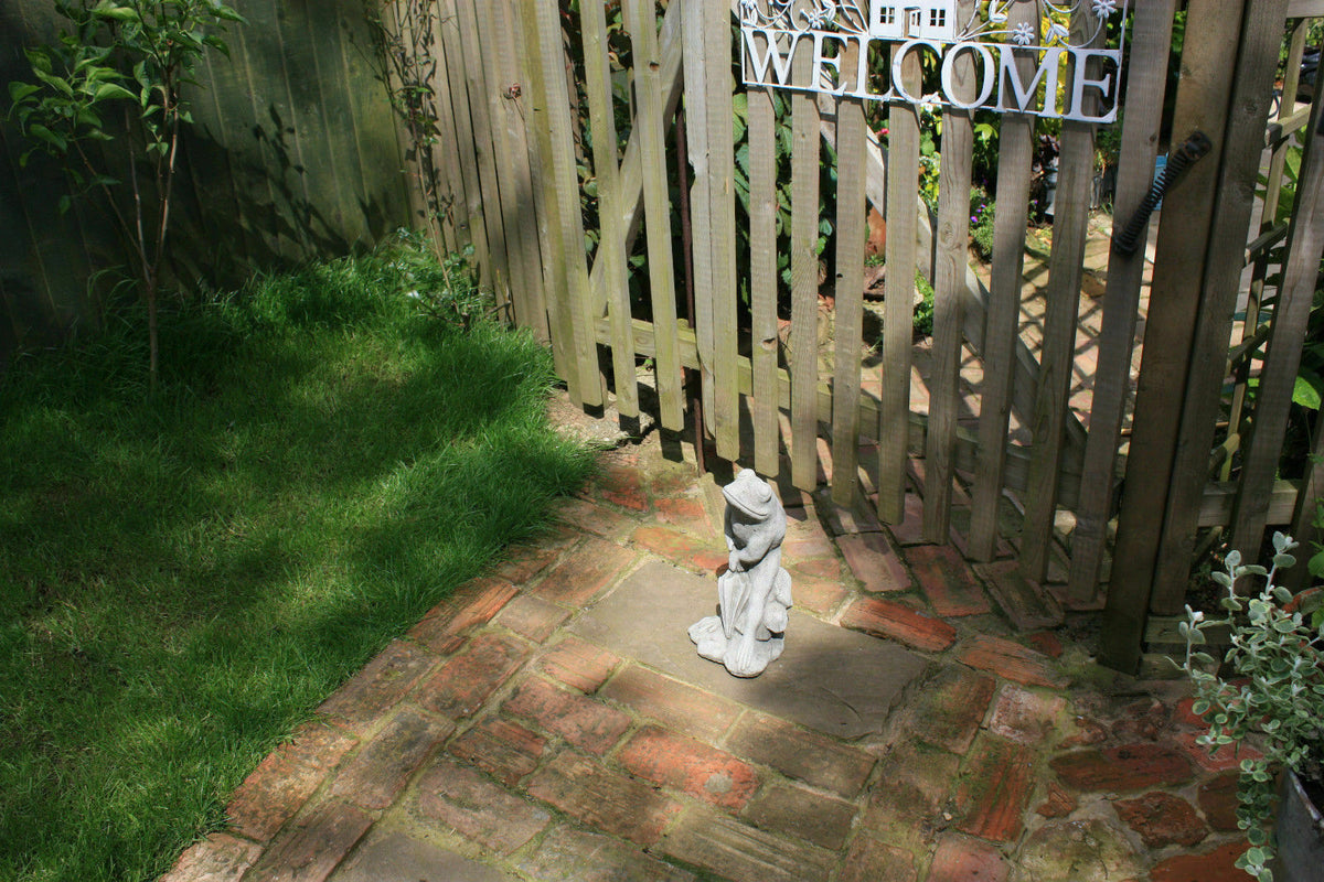 Stone Frog Garden Ornament with Umbrella placed on a brick path near a wooden gate, welcoming guests with unique garden charm.