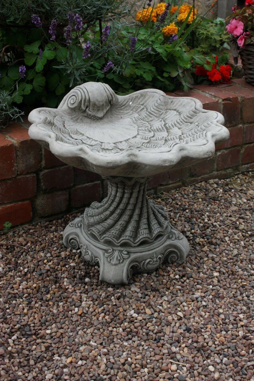 Stone Shell Bird Bath with elegant antique grey finish, set on gravel in a flower-filled garden. Ideal for outdoor decor and bird attraction.