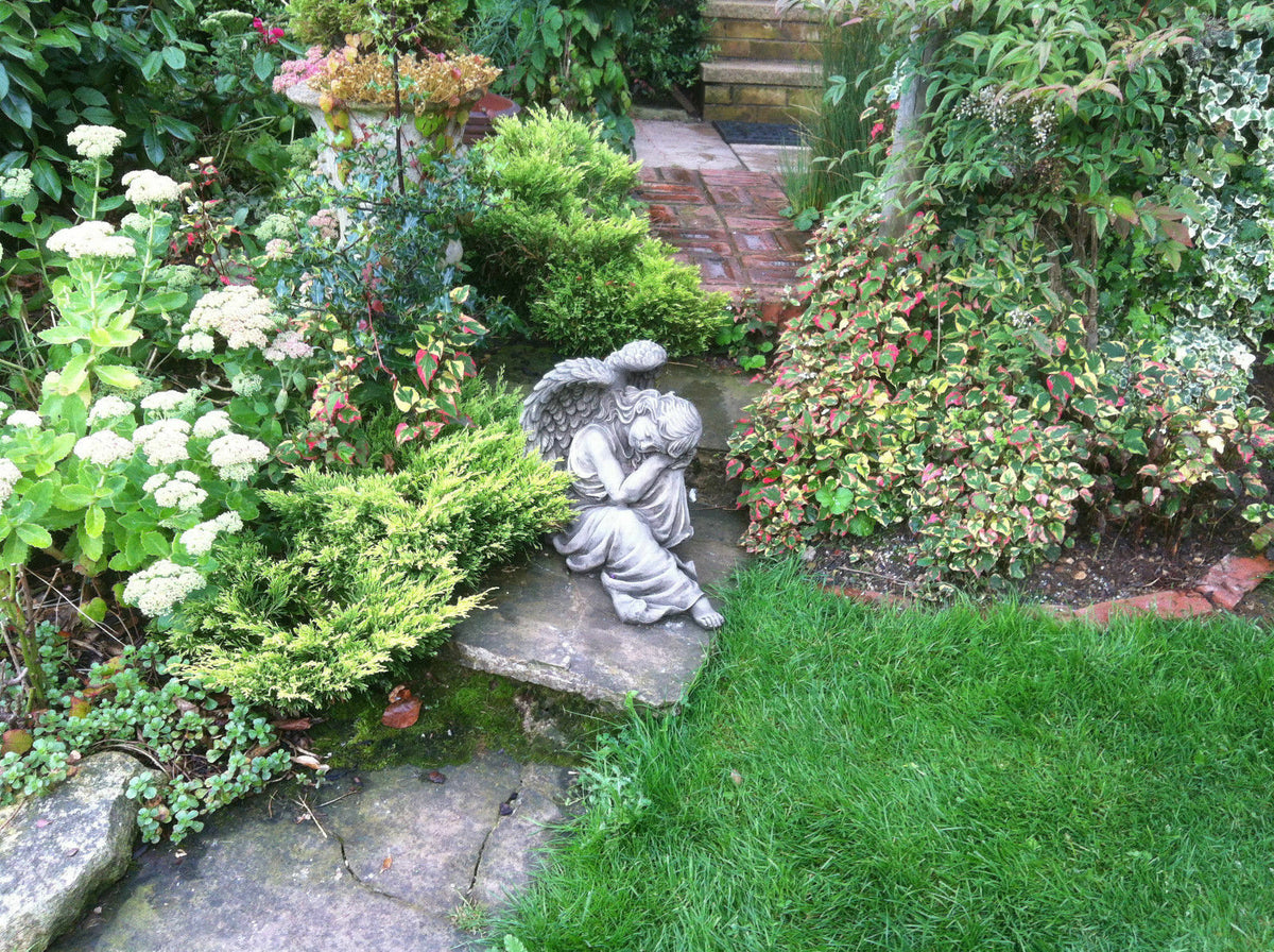 Stone Winged Angel Ornament nestled among lush garden plants on a stone slab, enhancing the natural landscape with serene charm.