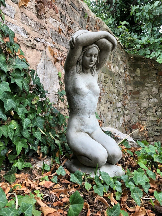 Kneeling Nude Lady Statue in antique grey stone, placed in a garden with ivy and stone wall backdrop. Ideal for outdoor decor and flowerbeds.