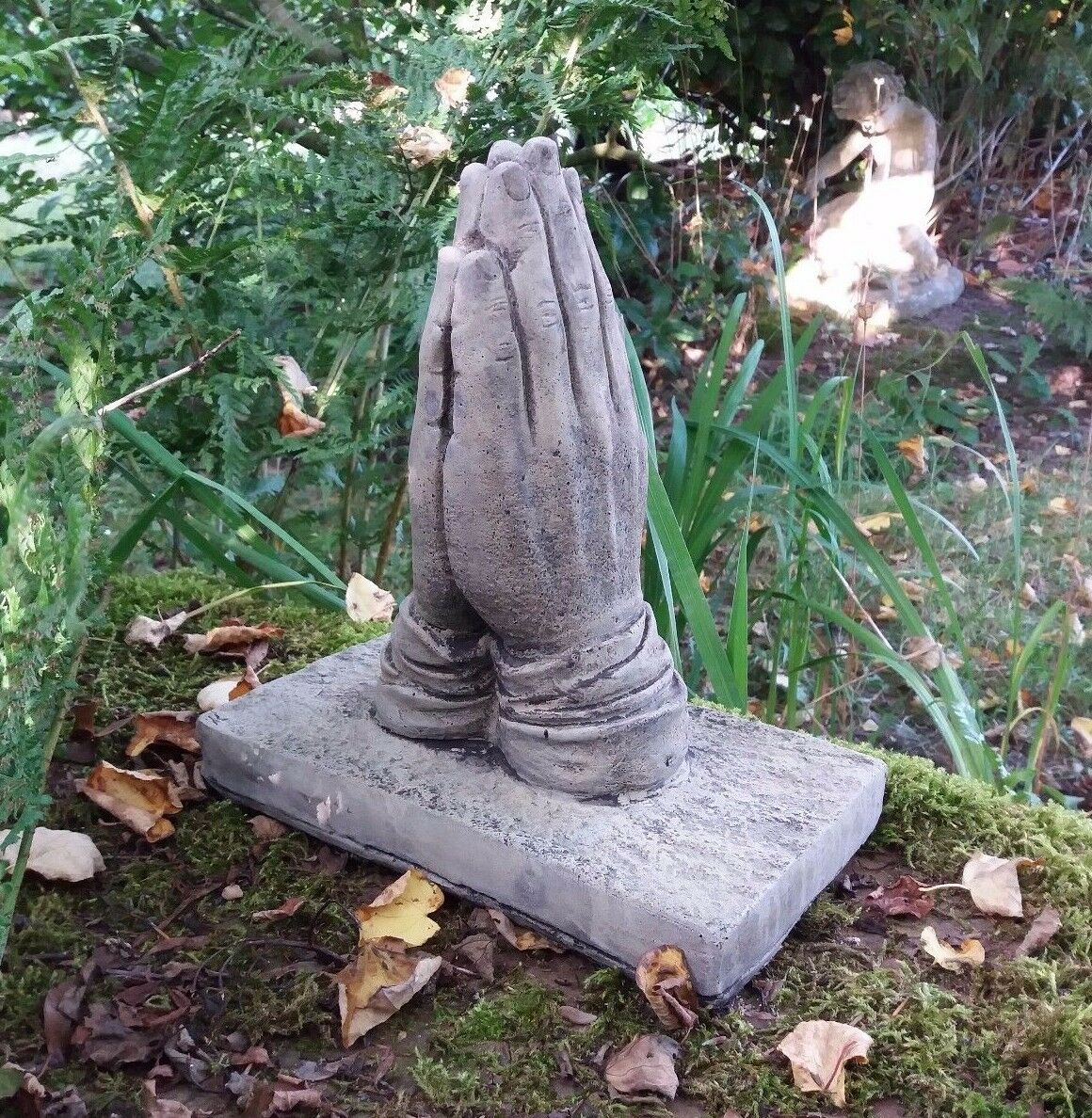 Memorial garden ornament of praying hands on mossy ground, handcrafted from stone. Frostproof and aged for outdoor durability.