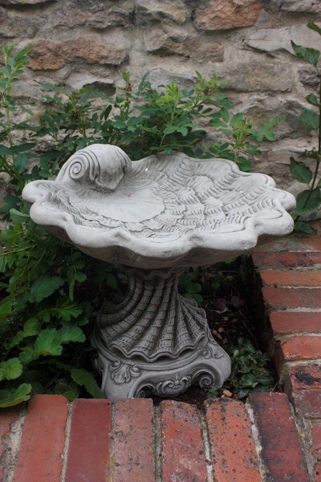 Large Stone Shell Design Bird Bath nestled in a brick garden corner, showcasing ornate detailing and durable stone craftsmanship.