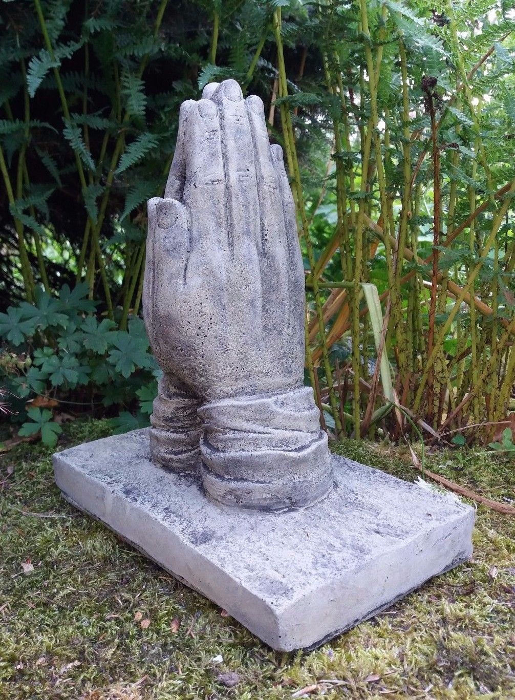 Outdoor stone sculpture of prayer hands on base, nestled in garden foliage. Handmade from crushed limestone with rustic finish.