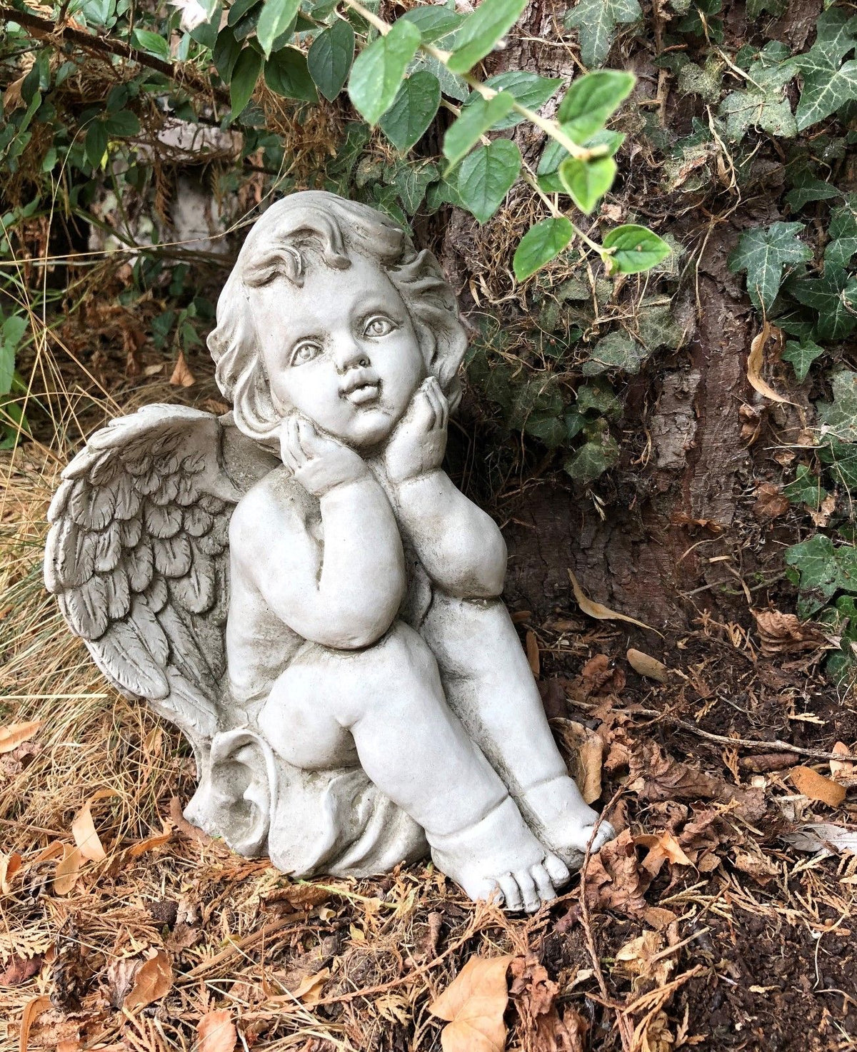 Thoughtful stone baby angel sculpture with hands on cheeks. Garden ornament adds charm to flowerbeds, porches, or memorials.
