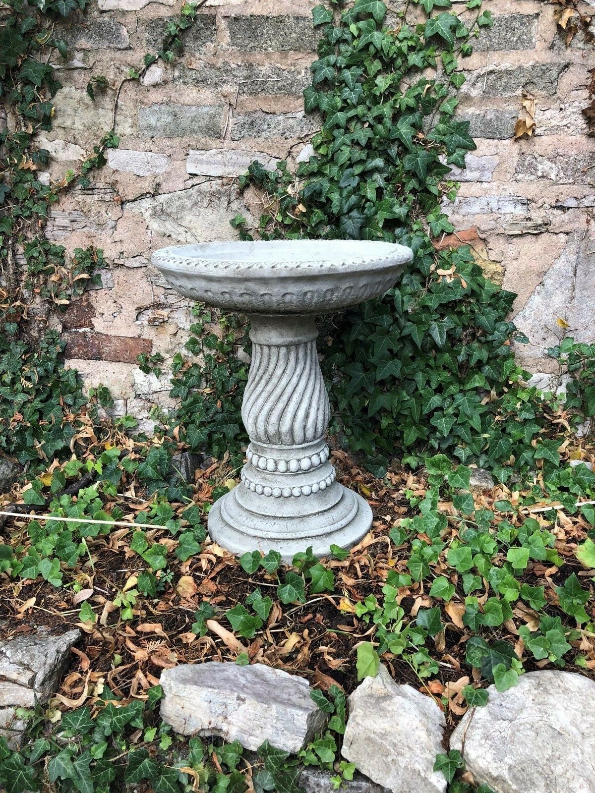 Classic stone garden bird bath with ornate twist base design nestled among ivy and rocks. Durable and stylish garden ornament.