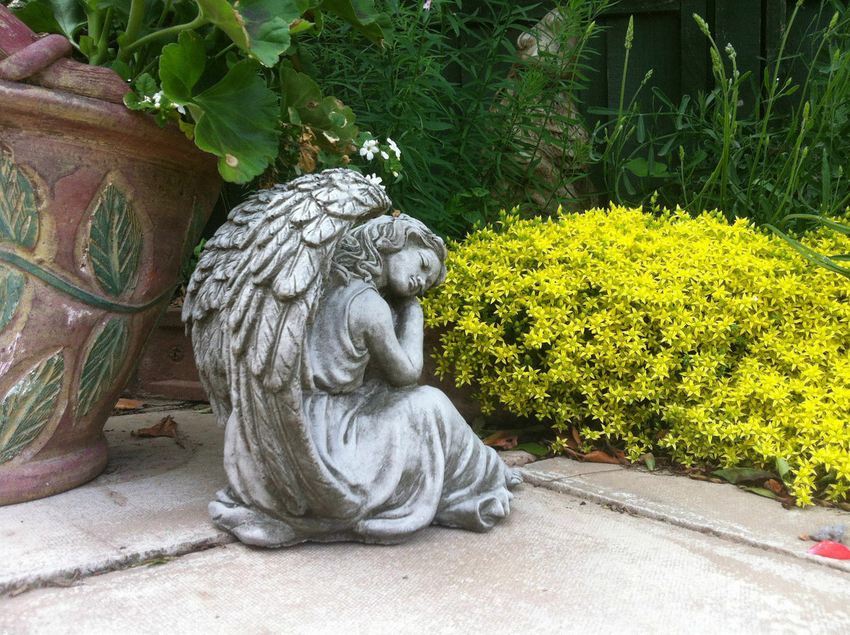 Grey stone angel sculpture with wings resting near yellow ground cover and potted plants, ideal for patios or flower beds.