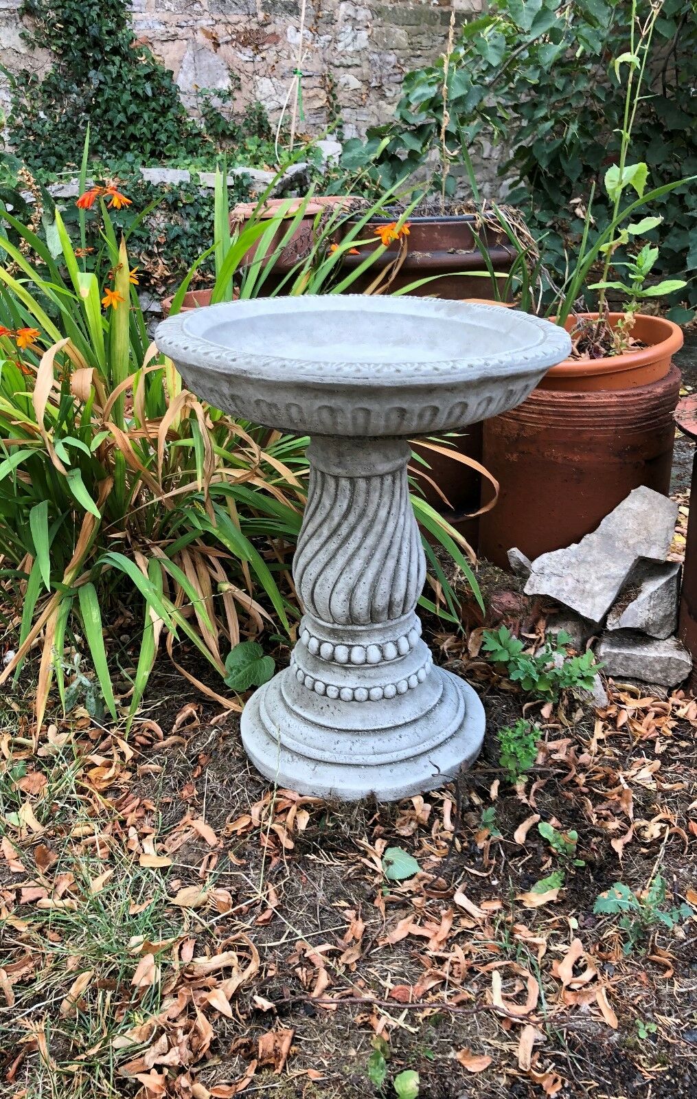 Handmade limestone bird bath with twisted base design, surrounded by potted plants and dry leaves. Ideal for decorative garden use.