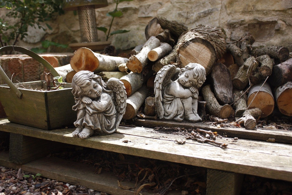 Pair of stone angel garden ornaments resting on a wooden pallet near stacked firewood. Durable decorative statues for outdoor decor.