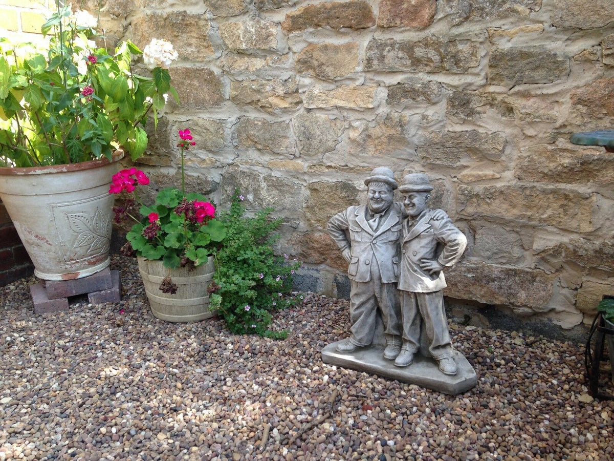 Full garden view featuring Laurel & Hardy stone statue as centerpiece among rustic stone wall, gravel, and flowering pots.