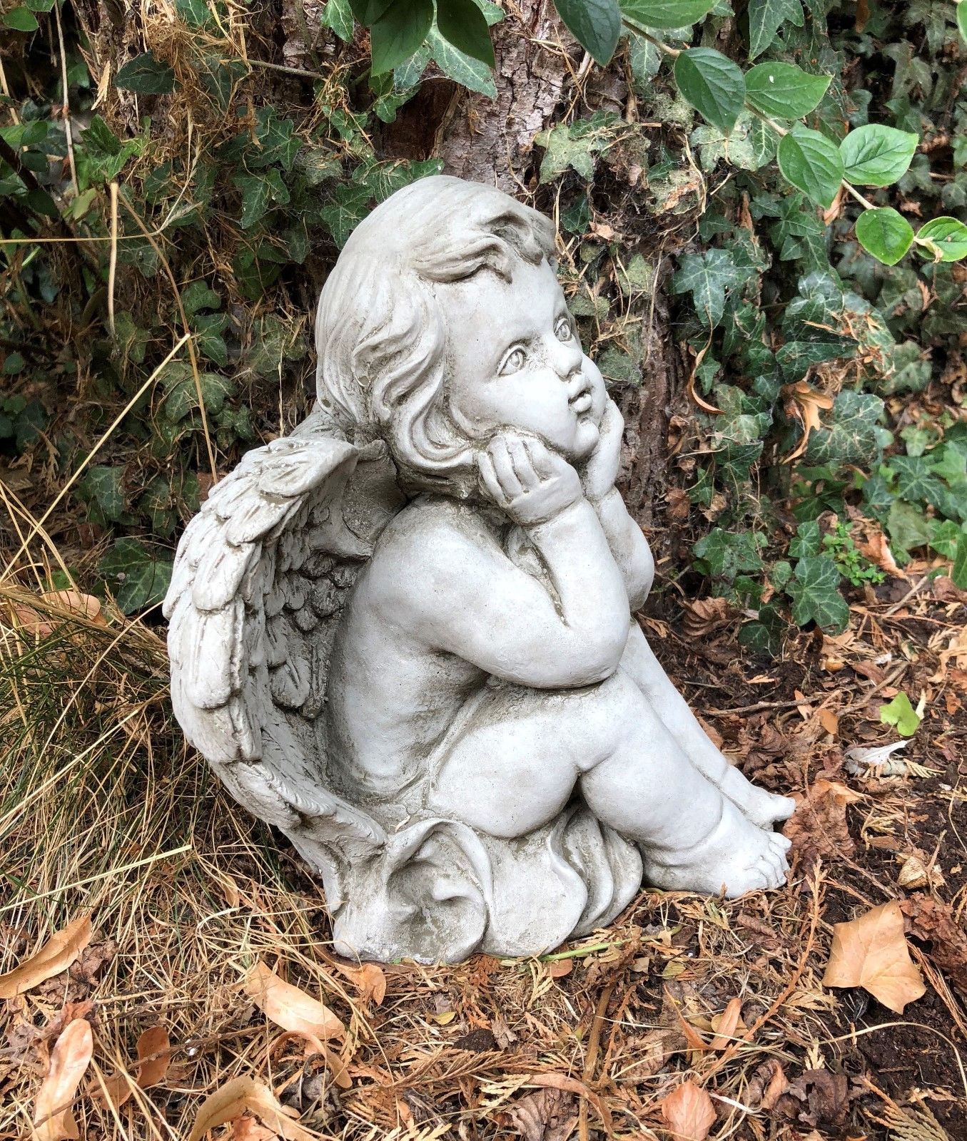 Side profile of stone winged baby angel sitting in reflective pose. Decorative garden sculpture for peaceful outdoor ambiance.