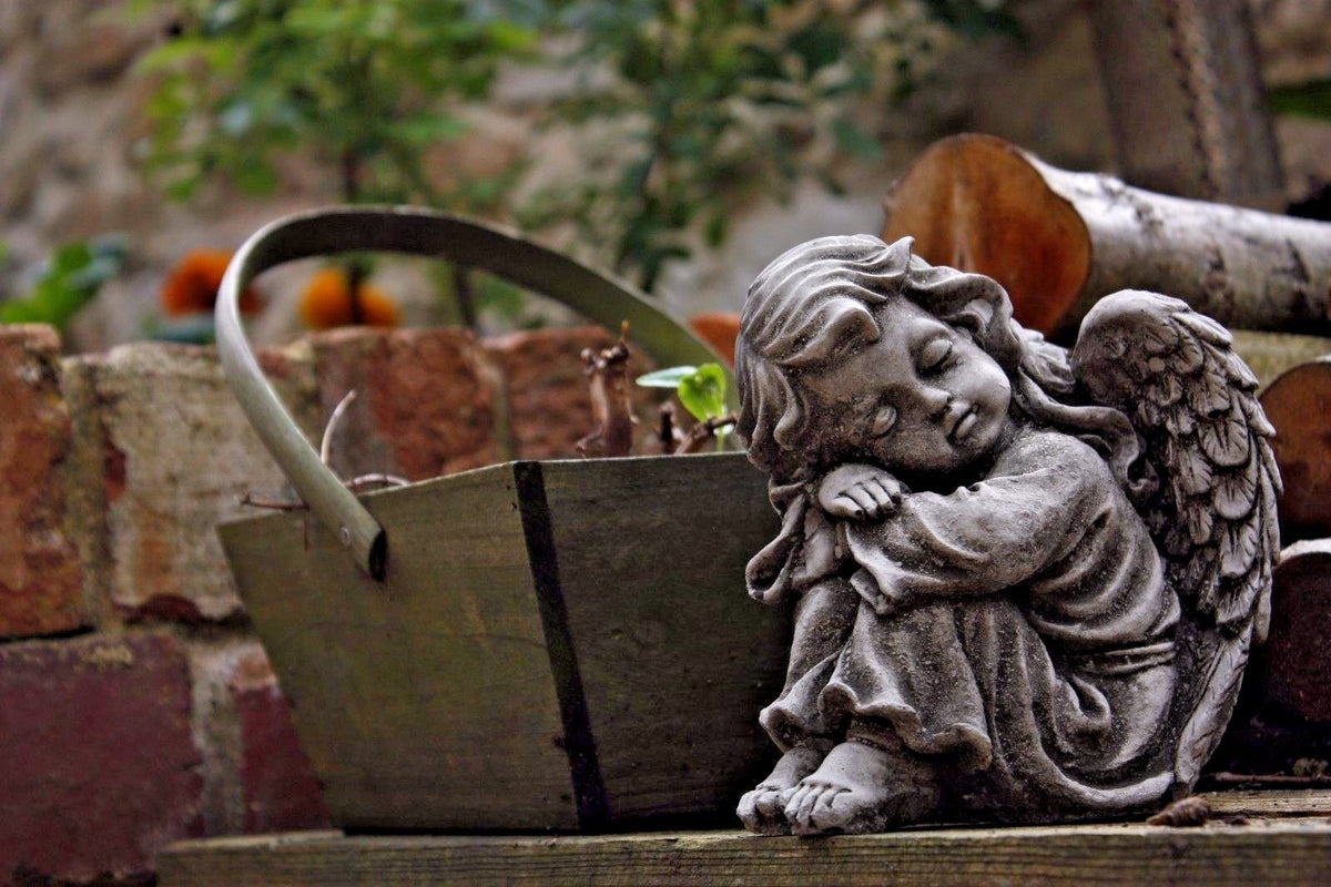 Close-up of a stone angel garden ornament beside a rustic wooden basket. Ideal for enhancing garden charm with durable limestone decor.