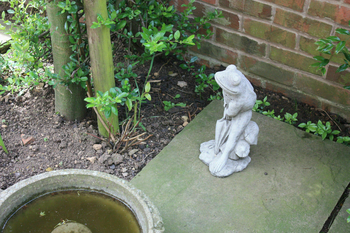 Stone Frog Garden Ornament with Umbrella placed beside a small pond and brick wall, enhancing garden ambiance with artistic flair.