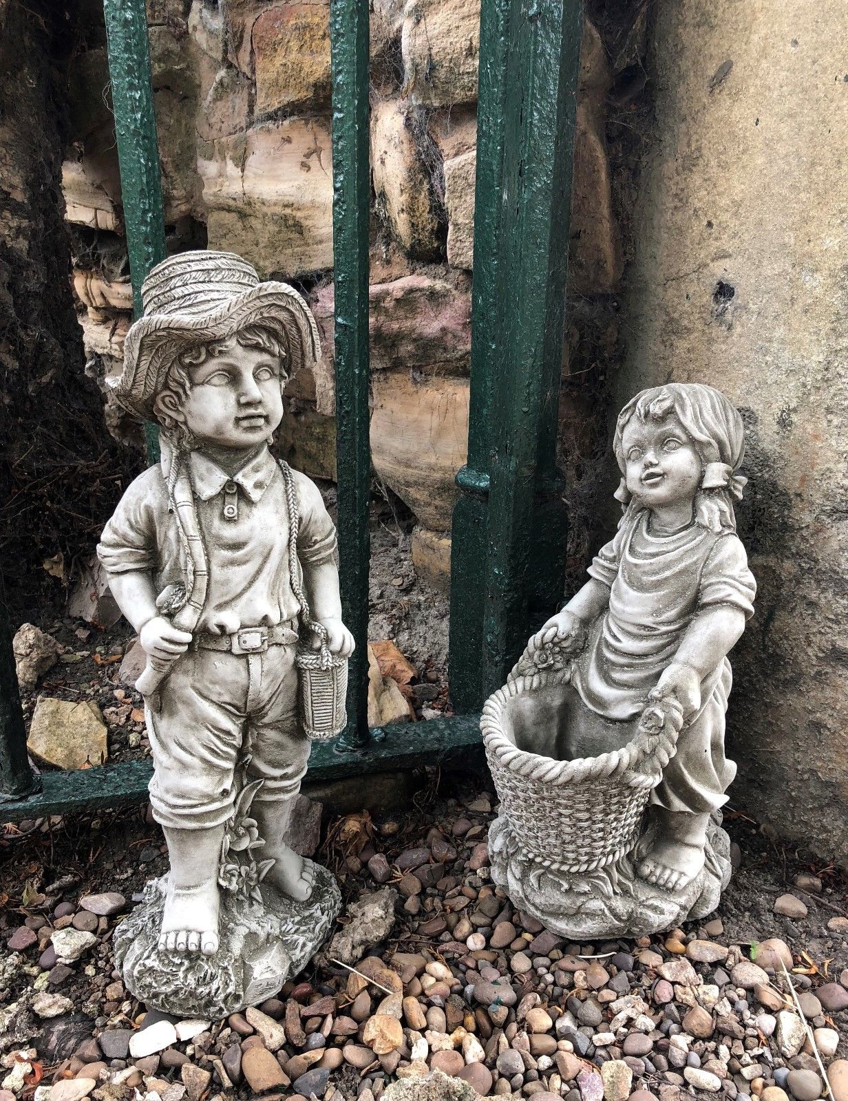 Garden stone ornaments of a fisher boy with a fishing rod and basket girl holding a planter, placed on gravel near a green iron fence.