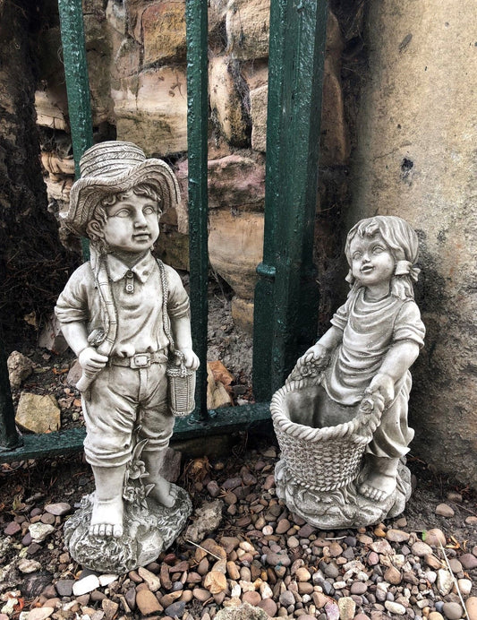 Garden stone ornaments of a fisher boy with a fishing rod and basket girl holding a planter, placed on gravel near a green iron fence.