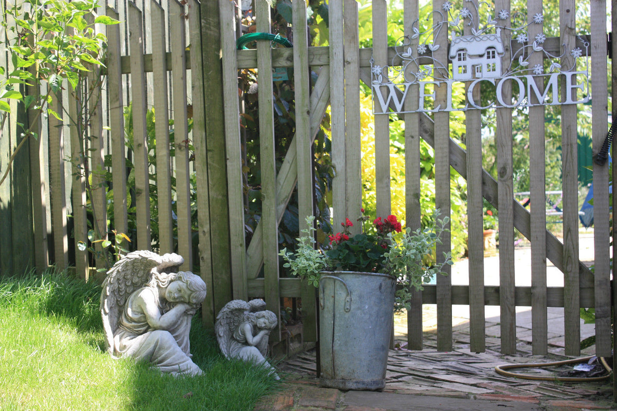 Pair of Grey Stone Winged Angel Ornaments by a wooden garden gate with welcome sign, adding a peaceful touch to outdoor entryways.
