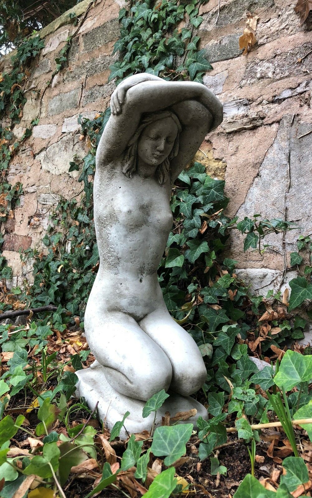 Detailed stone sculpture of a nude woman kneeling with arms overhead, ideal for enhancing garden aesthetics or outdoor art collections.