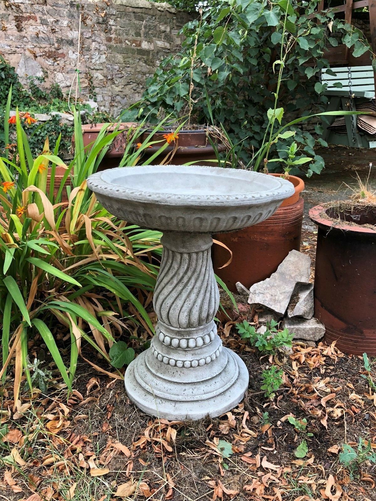 Stylish stone garden bird bath with twisted pedestal and wide bowl, set in a lush garden area. Great for small and large birds alike.