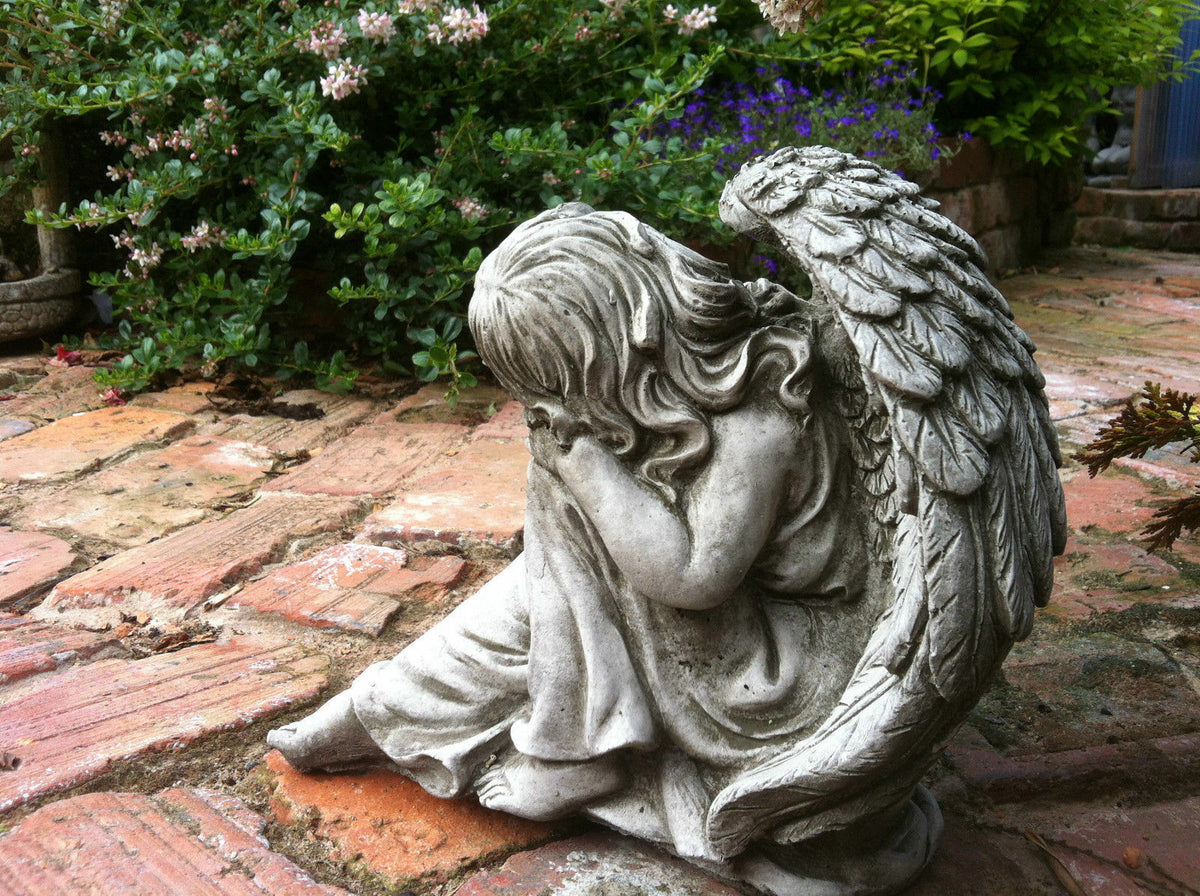 Durable Grey Stone Winged Angel Ornament seated on brick path, surrounded by green foliage, ideal for memorial or garden decor.