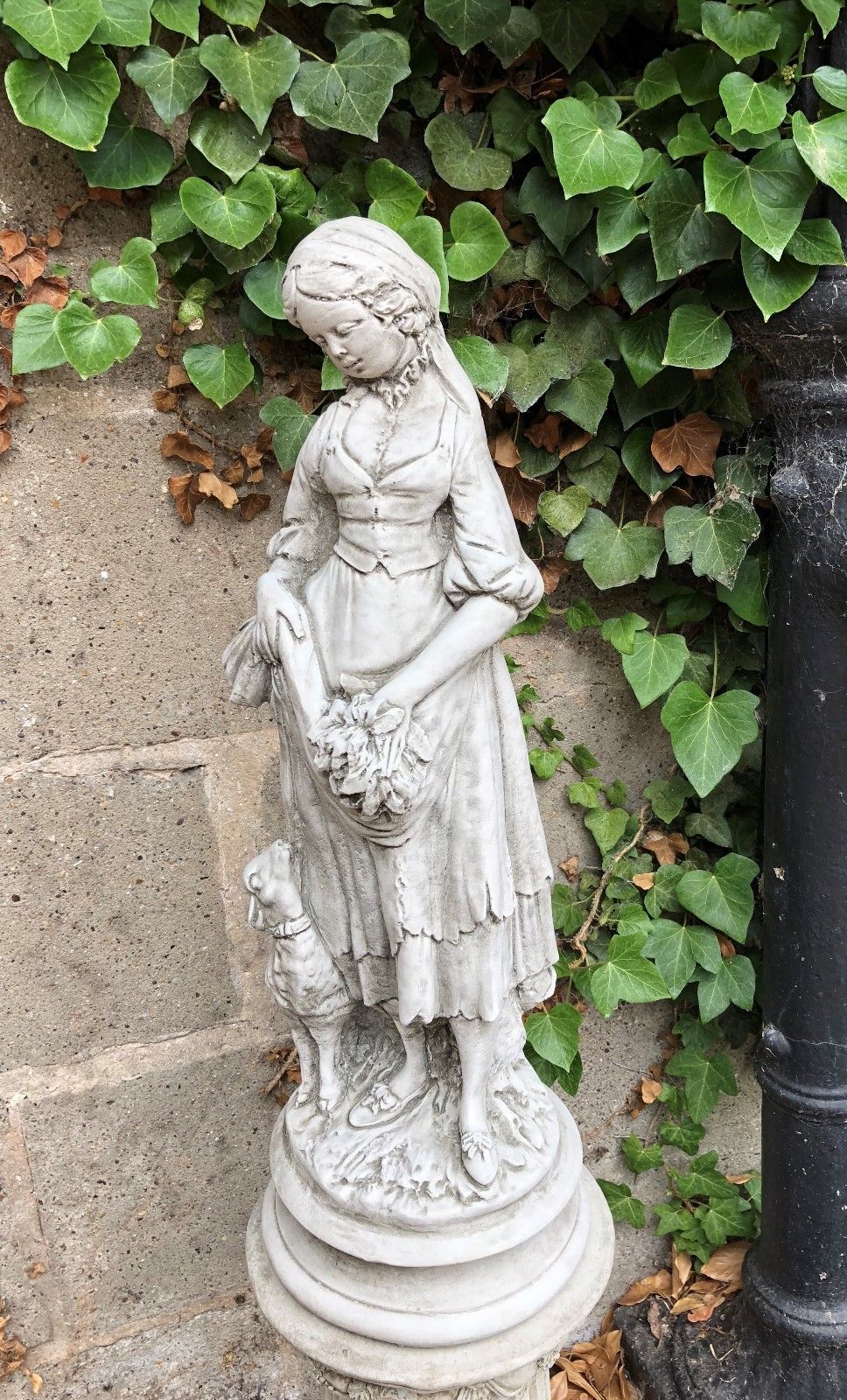 Side view of Stone Garden Shepherdess Statue in rustic dress, holding flowers, perfect for enhancing garden aesthetics with vintage charm.