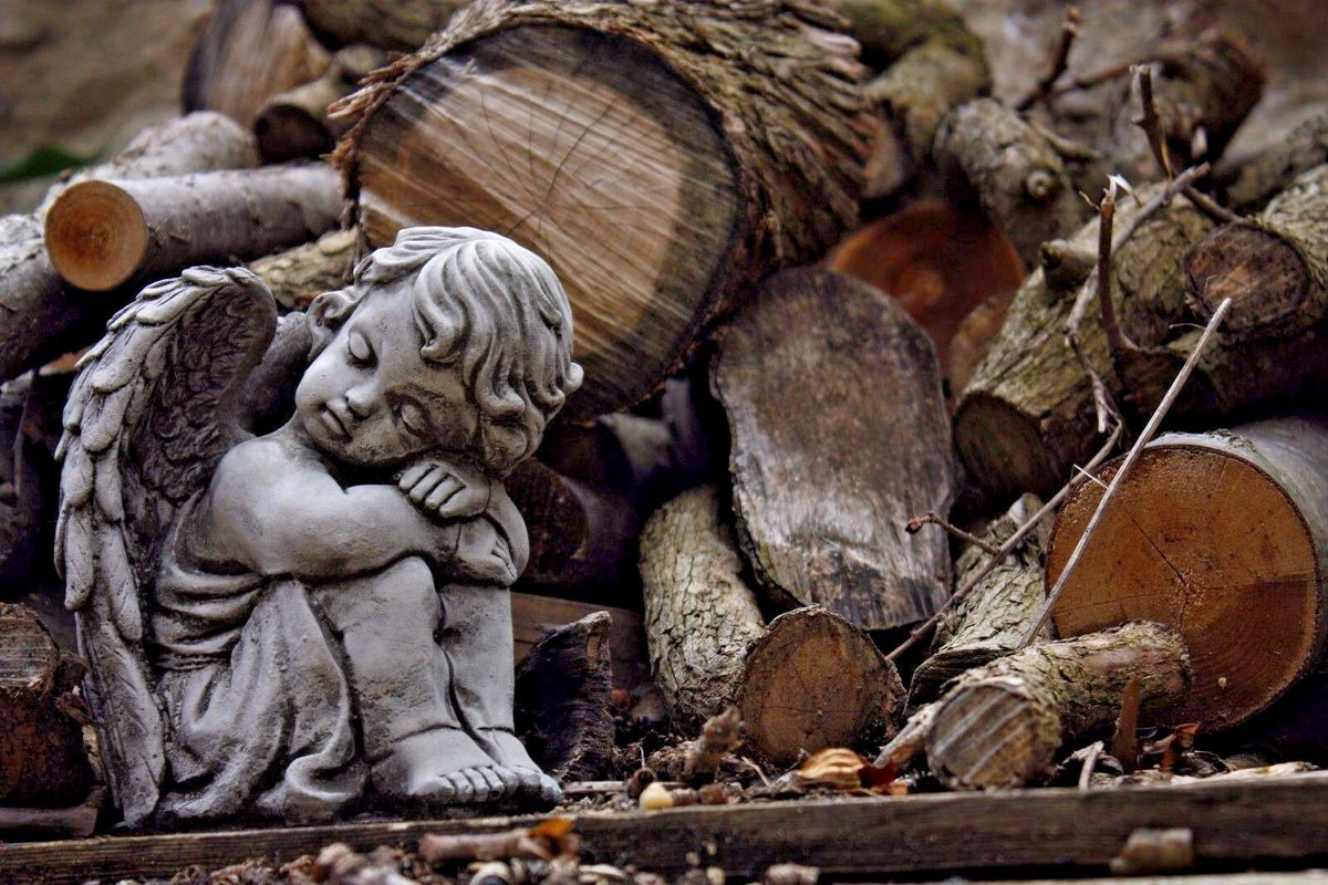 Single stone angel garden ornament resting near chopped firewood. Durable limestone statue adds a peaceful touch to garden decor.