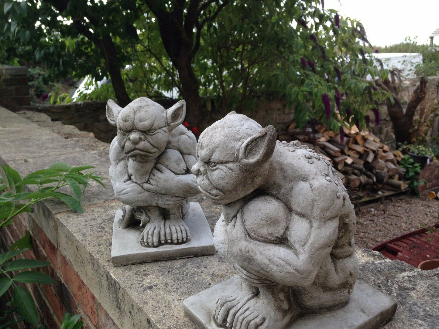 Pair of Grumpy Stone Gargoyle Statues on garden wall with greenery, perfect for gothic outdoor decor. Weatherproof limestone garden ornaments.