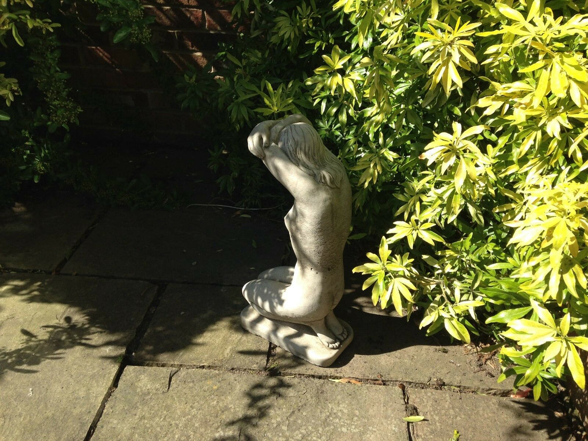 Stone nude lady statue kneeling on patio surrounded by greenery. Weather-resistant garden ornament for UK outdoor spaces.