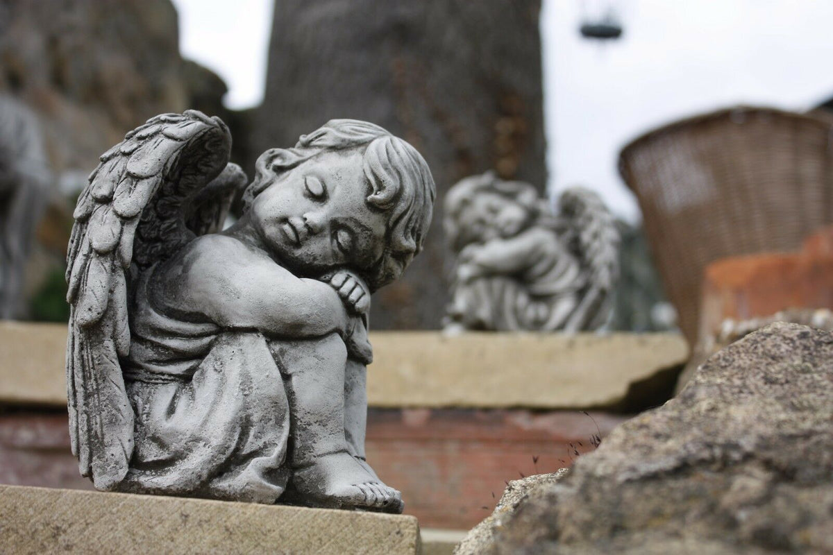 Stone angel garden ornament sitting on a stone ledge with another in the background. Perfect for patios and flowerbeds with timeless charm.