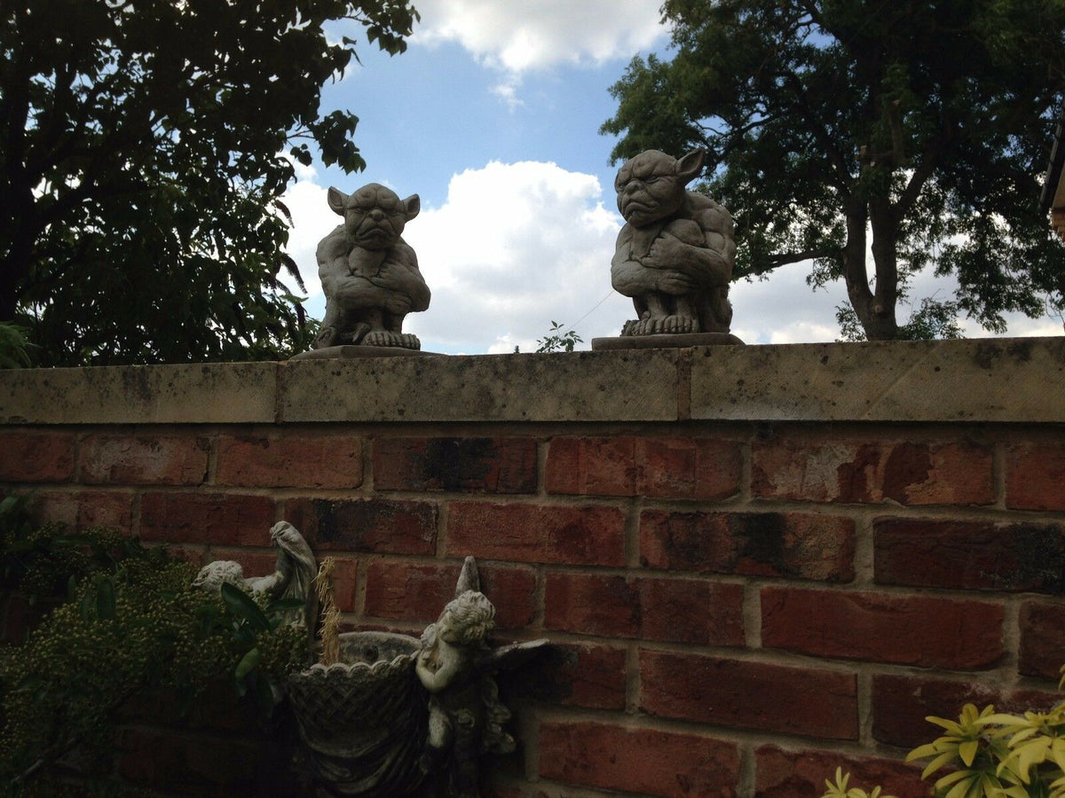 Grumpy Stone Gargoyle pair on red brick wall in garden setting. Gothic lawn ornaments for outdoor decor and landscaping accents.