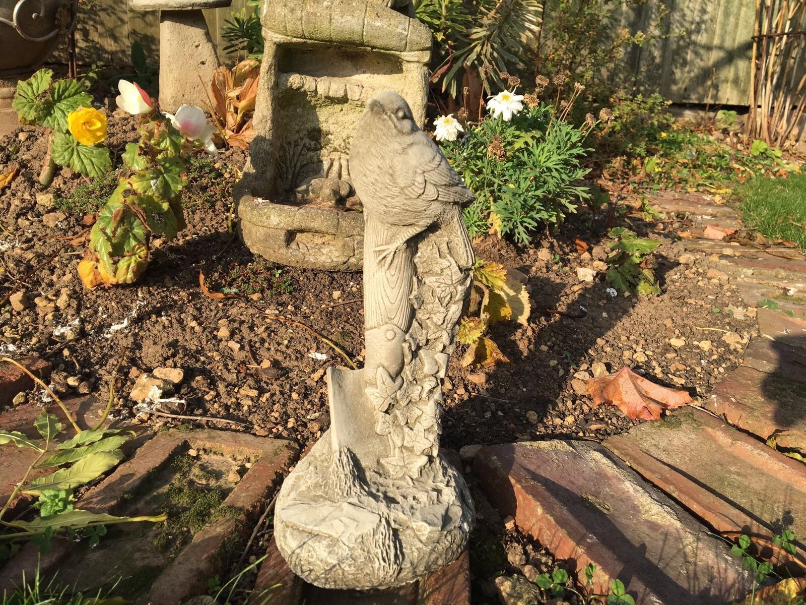 Stone Bird Garden Ornament on Spade with ivy detail, placed in a flower bed. Durable limestone sculpture for outdoor garden decor.