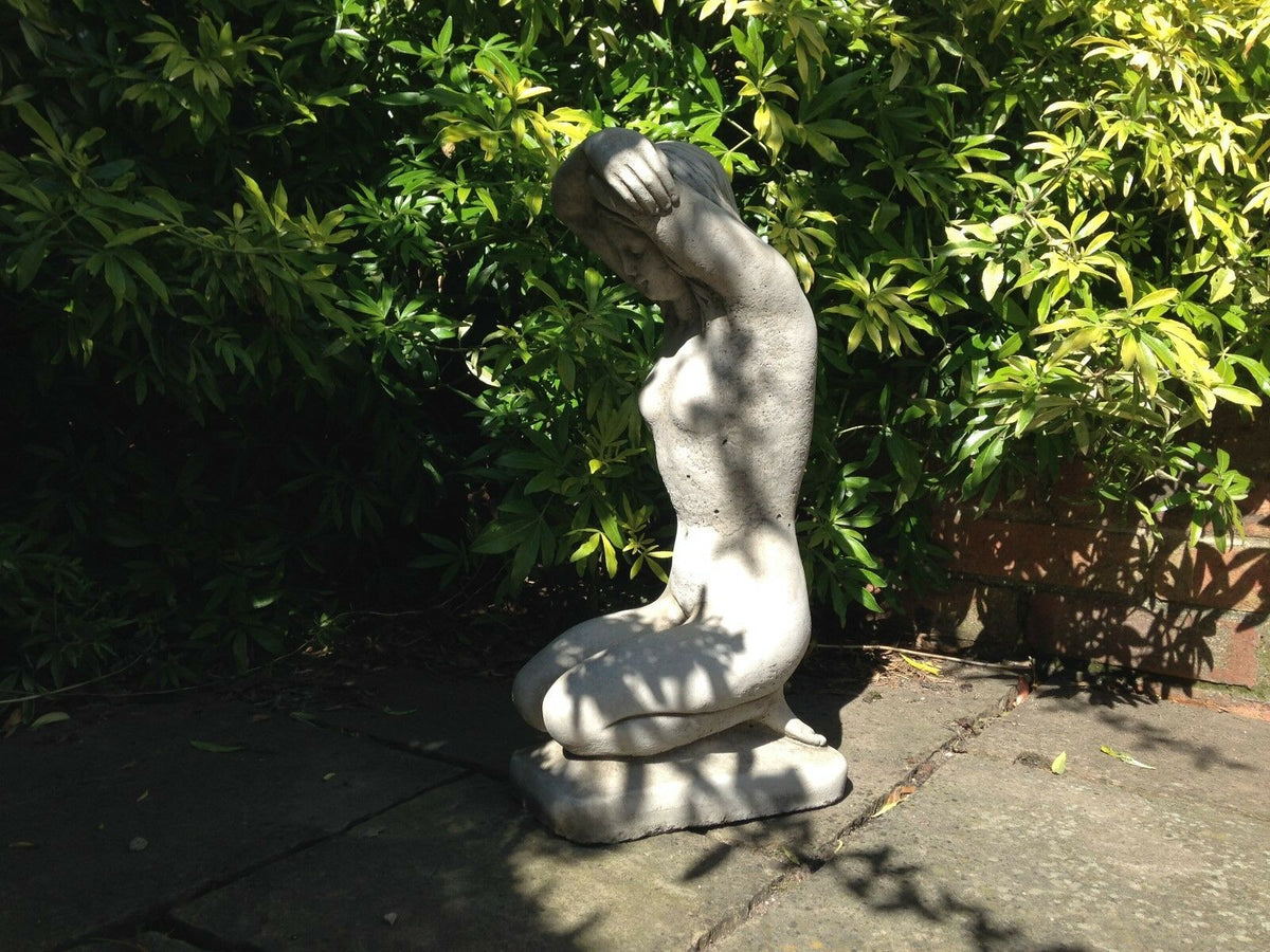 Kneeling Nude Lady Garden Sculpture in limestone, partially shaded by foliage. Ideal for patios, porches, or landscaped gardens.