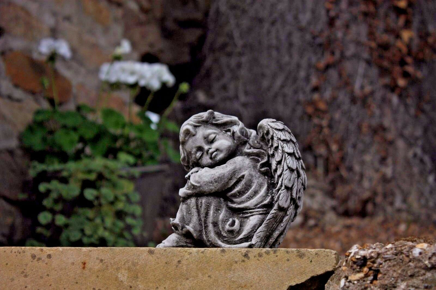 Stone angel garden ornament leaning peacefully on a stone wall with greenery behind. Handcrafted outdoor statue for serene garden ambiance.